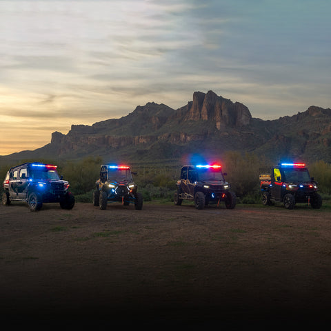 Four off-road vehicles with lights on in a desert landscape at dusk.